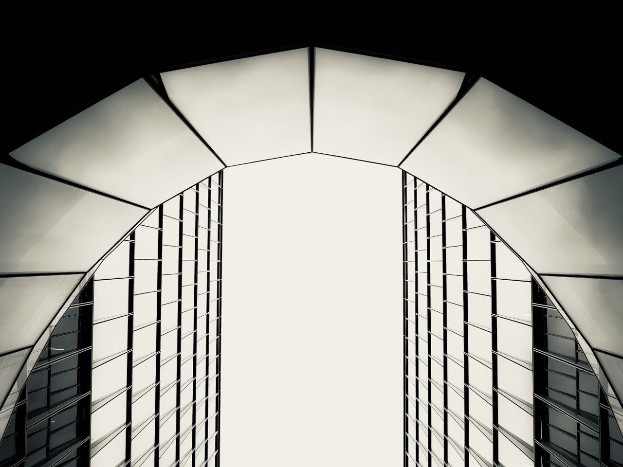 Looking up at an arched glass-and-steel building against a pale sky, in black and white
