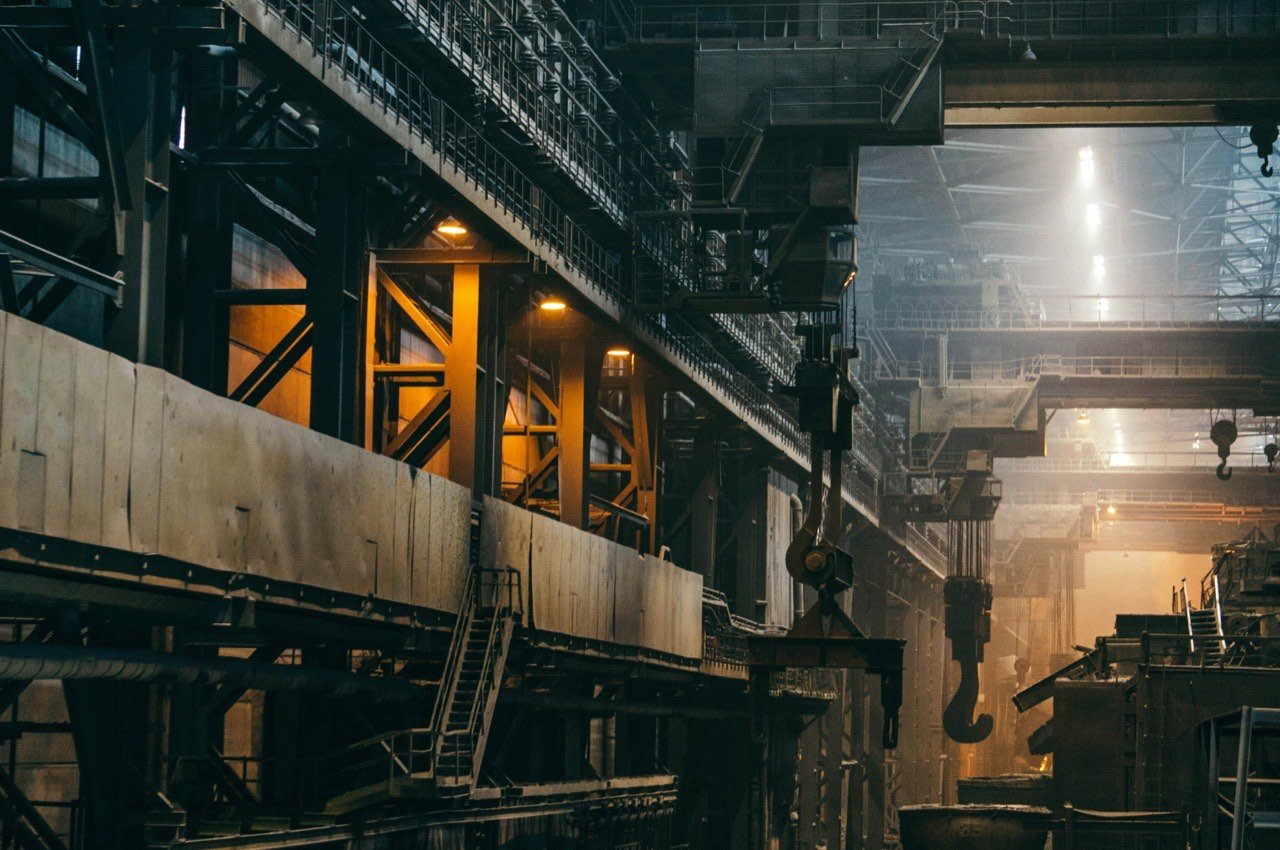 The inside of a large industrial hall with overhead cranes, hooks and steel walkways, lit by shafts of light