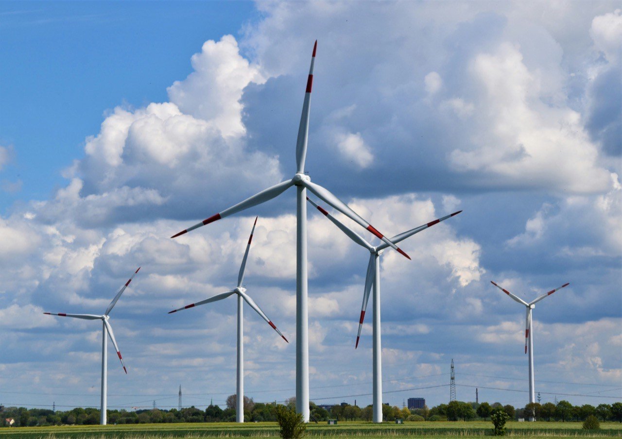 Five white wind turbines with red-tipped blades on a green field under a cloudy blue sky